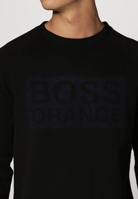 Sudadera negra con un gráfico texturizado que dice "BOSS ORANGE" en azul oscuro. Presenta un cuello redondo y mangas raglán. Tejido suave.