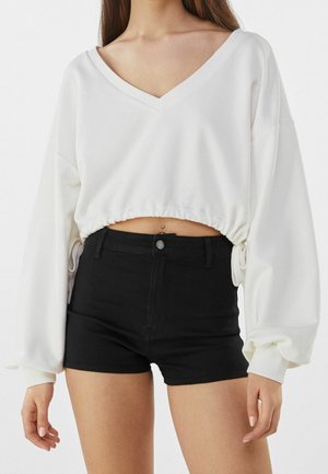 Kvinde iført en hvid, kort V-hals top med lange ærmer og snøre samt sorte, højtaljede shorts, der viser en navlepiercing.