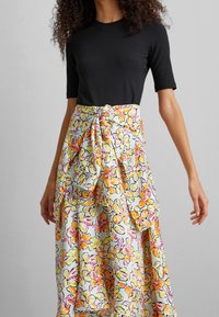 Jupe fleurie avec ceinture à nouer en tissu léger. Présente des fleurs vives orange, jaune et rose sur fond blanc. Haut noir à manches courtes.