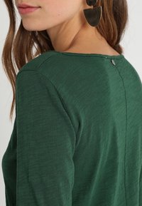 Green garment with a soft texture, featuring a back button detail and a rounded neckline, highlighting a simple and clean design.