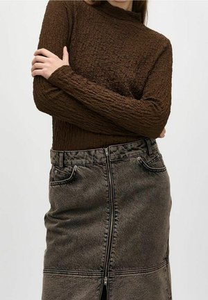 Person iført en brun, tekstureret langærmet top, der er stoppet ned i et højtaljet sort denimskørt med en central lynlås.