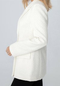 Bílý strukturovaný blazer s přiléhavým střihem, klopovými límci a boční kapsou. Materiál má tkaný vzor pro zvýraznění detailů.