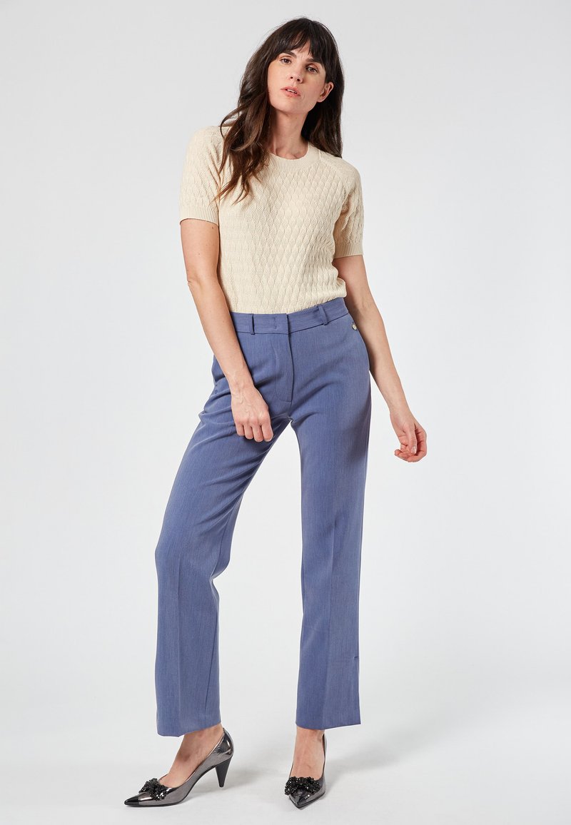 Cream knitted short-sleeve top, light blue tailored trousers, and black pointed-toe heels with decorative accents. Simple, structured design.