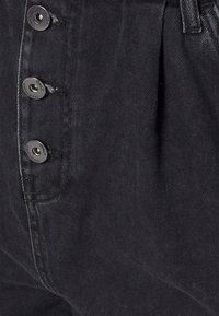 Mustat denimpantsit, joissa on vetoketju ja neljä suurta metallipainiketta. Ominaisuuksiin kuuluvat näkyvät ompeleet ja sivuhelmat lisätilavuuden lisäämiseksi.