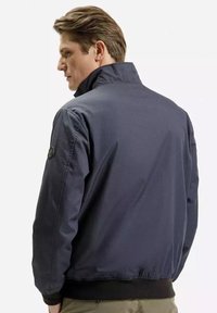 Granatowa kurtka bomberka, wykonana z gładkiego materiału, charakteryzuje się wysokim kołnierzem i elastycznym dołem. Zawiera detal z naszywką na lewym rękawie.
