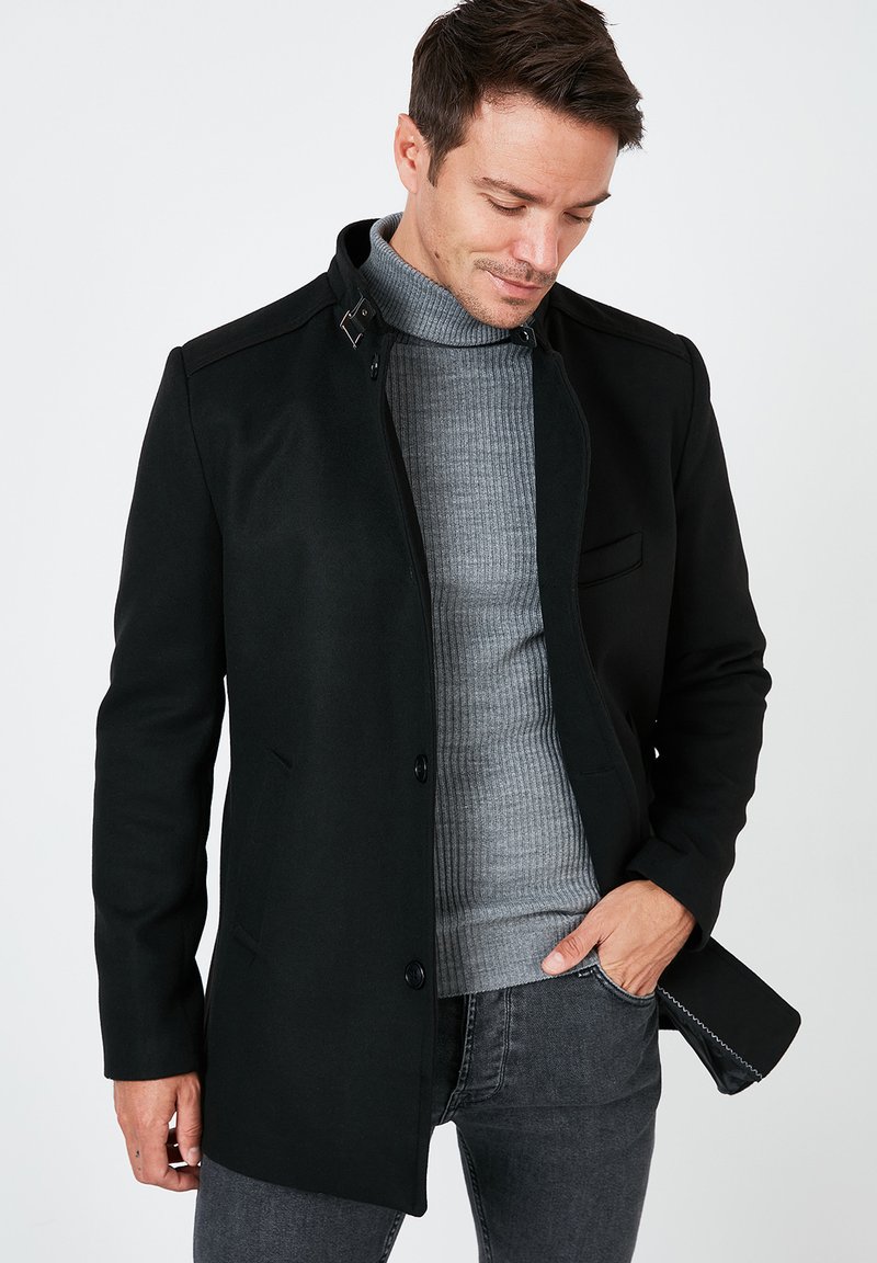 Schwarzer Wollmantel mit hohem Kragen, Knopfverschluss und Fronttaschen, getragen über einem grauen gerippten Pullover. Glatte Textur mit taillierter Passform.