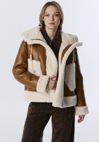 Bruine leren jas met crème schapenvacht voering, capuchon en voorzakken. Voorzien van een ritsluiting en verstelbaar trekkoord.