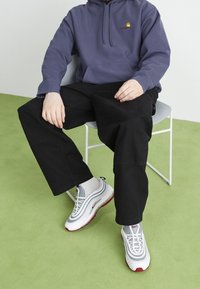 Osoba siedząca na białym krześle, ubrana w fioletową bluzę Carhartt, czarne spodnie i białe sneakersy Nike, na zielonym wykładzinie.
