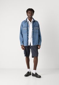 Veste en jean avec deux poches poitrine, chemise blanche en dessous, shorts foncés et mocassins noirs avec des chaussettes blanches.