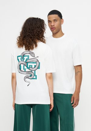 Deux mannequins portent des t-shirts blancs et des pantalons verts, l’un montre un graphisme dans le dos avec un serpent enroulé au-dessus de grandes lettres bleu sarcelle.
