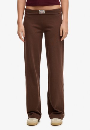 Pantalon marron à jambes larges avec un patch logo Superdry sur la ceinture, porté avec des baskets beige et un haut court marron rouge.