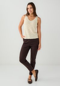 Jasno beżowy bezrękawnik z dekoltem w kształcie litery V, zestawiony z ciemno brązowymi spodniami slim-fit. Modelka nosi czarne płaskie buty w siateczkę.