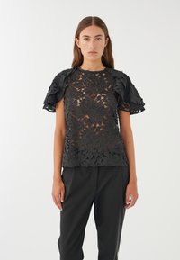 Top in pizzo floreale nero con maniche corte a volant, caratterizzato da un design trasparente e un colletto rotondo, abbinato a pantaloni neri sartoriali.