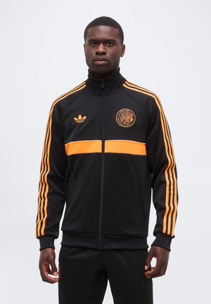Άνδρας που φοράει μαύρο αθλητικό μπουφάν Adidas με πορτοκαλί ρίγες, πορτοκαλί οριζόντιο λουρί και λογότυπα στο στήθος και στα παντελόνια, στέκεται μπροστά σε λευκό φόντο.