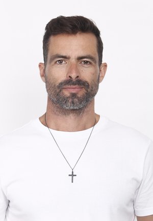 Men's silver cross pendant necklace on a thin chain, worn over a plain white t-shirt. Hair is dark and slightly tousled.