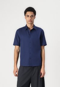 Camisa de manga corta azul marino hecha de una tela suave, con un cierre frontal de botones y un cuello clásico. Combinada con pantalones grises holgados.