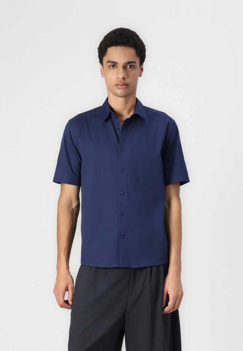 Camisa de manga corta azul marino hecha de una tela suave, con un cierre frontal de botones y un cuello clásico. Combinada con pantalones grises holgados.