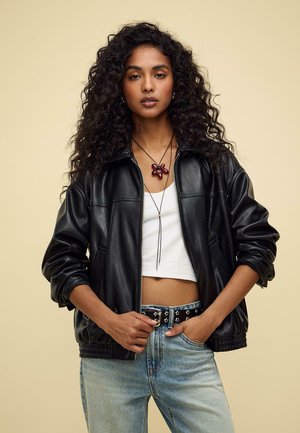 Schwarze Lederjacke mit Reißverschluss und lässiger Passform, kombiniert mit einem weißen Croptop und hellblauen Jeans. Auffälliges rotes Blumenhalsketten-Detail.