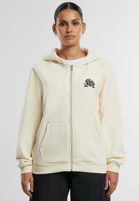 Cremfarbene Zip-Hoodie aus weichem Stoff. Mit einem schwarzen Rosendesign auf der linken Brust und Kängurutaschen. Mit gerippten Bündchen und Saum.
