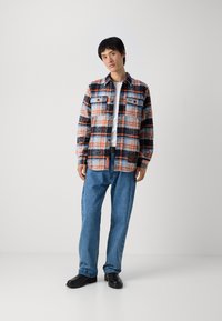 Camicia di flanella a quadri in arancione, blu e nero, con due tasche sul petto, design con bottoni, indossata sopra una maglietta bianca e jeans blu.