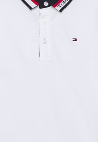 Valkoinen polo-paita, jossa on tummansininen kaulus, jossa on punaisia ja valkoisia raitoja. Kolme nappia napituksessa. Pieni logo yksityiskohta vasemmassa rinnassa.