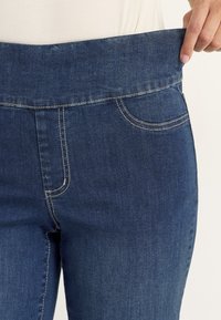 Persoon die de tailleband van een blauwe spijkerbroek van het lichaam wegtrekt om de losheid te tonen, draagt een lichtgekleurde top, op een neutrale achtergrond.