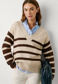 Beżowy sweter dzianinowy z szerokimi brązowymi paskami, dekoltem w kształcie litery V i ściągaczem. Nosi go na jasnoniebieskiej koszuli z falbanami. Trzyma ciemnoniebieską torebkę.