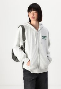 Harmaa vetoketjullinen huppari, jossa on nauhavetoketju, vihreä brodeerattu logo ja etutaskut. Yhdistetty tummiin farkkuihin ja mustaan laukkuun.