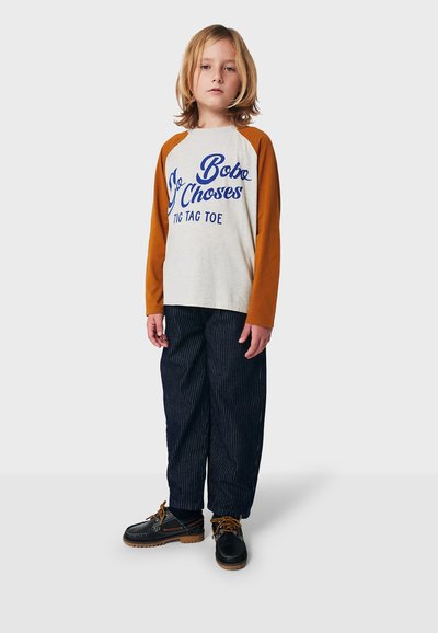 Langärmliges Raglan-T-Shirt mit grauem Körper und rostfarbenen Ärmeln, mit blauem Grafiktext. Kombiniert mit gestreiften dunklen Hosen und schwarzen Schuhen.