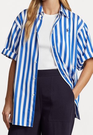 Vrouw met een blauw-wit gestreept overhemd met verticale strepen, gedragen over een wit tanktop, gecombineerd met een donkere broek, hand in de zak.