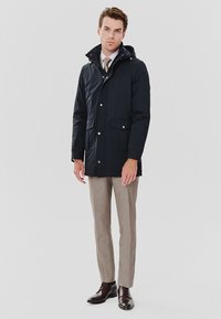Granatowa parka z kapturem, z napami i wieloma kieszeniami. Noszona na jasne, paskowane spodnie oraz brązowe buty.