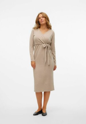 Beiges, langärmliges Kleid mit einem Wickeldesign und Taillenschnürung. Sanfte Textur, knielang und mit V-Ausschnitt. Getragen mit schwarzen Ballerinas.