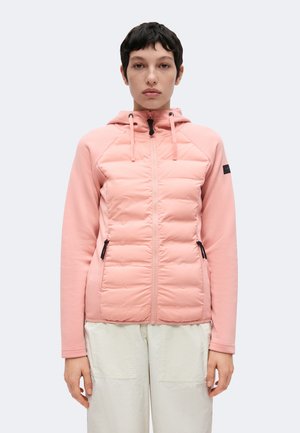Personne aux cheveux courts et foncés porte une veste à capuche rose clair zippée et un pantalon clair, debout devant un fond uni clair.