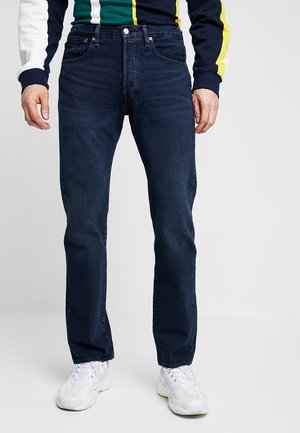Man draagt donkerblauwe rechte spijkerbroek, witte sneakers en een longsleeve met groene, gele, marineblauwe en witte strepen.