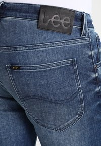 Pantalones vaqueros de mezclilla azul con un parche de cuero oscuro que lleva el logotipo "Lee", costuras visibles y un diseño clásico de bolsillo con un ligero efecto de desgaste.