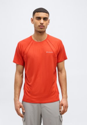 Vyras dėvi raudoną trumpomis rankovėmis Columbia marškinėlį ir šviesiai pilkas krovinių kelnes, stovintis prieš paprastą šviesią foną.