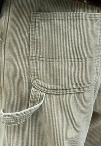 Beige kordfløyelstoff med vertikale riller, som har en sidelomme og søm-detaljer. Bukseben med en strukturert overflate.