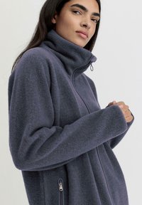 Giacca in pile viola scuro con colletto alto, chiusura frontale con zip e tasche laterali dotate di cerniere argento; texture morbida e vestibilità comoda.