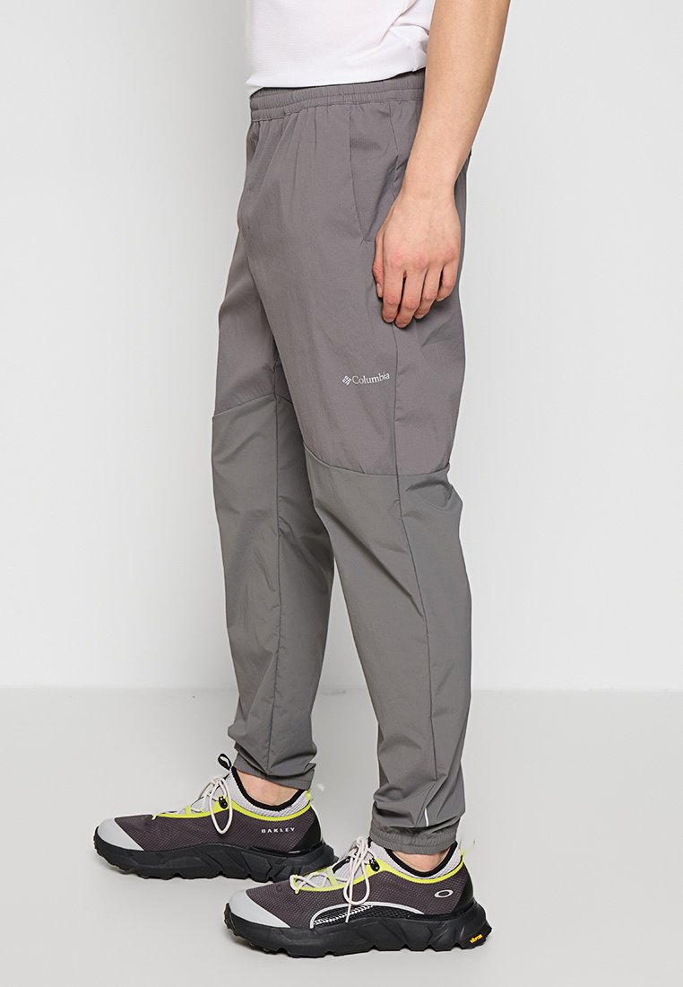 Mężczyzna w dwukolorowych, szarych spodniach Columbia i czarnych sneakersach Oakley, stojący na jednolitym, jasnoszarym tle.