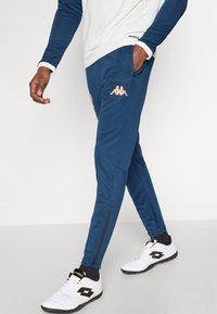 Μπλε ναυτικού χρώματος τζόγκερ με λεπτή εφαρμογή, διαθέτουν κεντημένο λογότυπο σε ροδακινί, τσέπες με φερμουάρ και αντίθετες μαύρες λεπτομέρειες κοντά στους αστραγάλους.