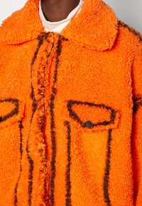 Oranžová fleecová bunda s texturovaným povrchem, černým ozdobným šitím a dvěma předními kapsami se zapínáním na patentky.