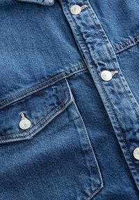 Camicetta in denim blu scuro con una texture leggermente sbiadita, dotata di bottoni a pressione argentati e due tasche sul petto con alette appuntite.