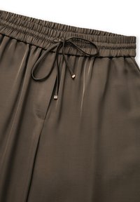 Bruine broek met elastische tailleband en trekkoordsluiting, met een gladde textuur, plooien en gouden accenten op de uiteinden van het trekkoord.