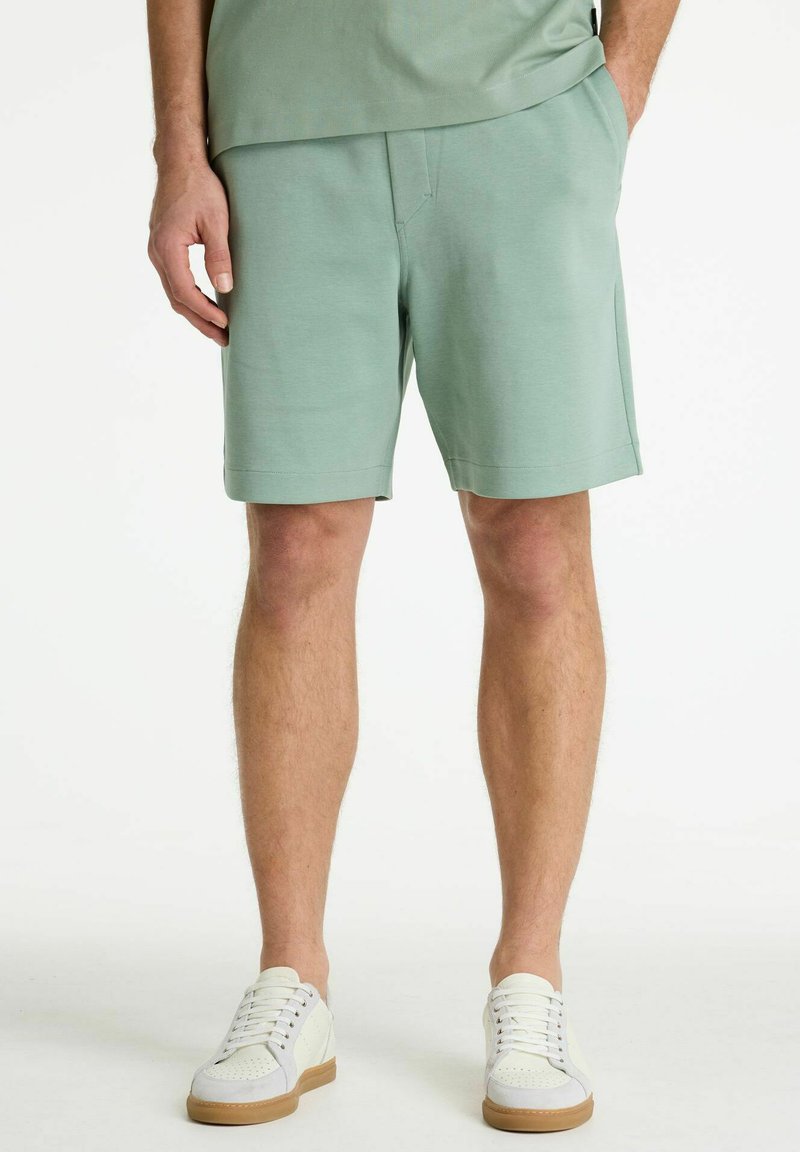 Man met lichtgroene korte broek en witte sneakers, staand met één hand in de zak tegen een witte achtergrond.