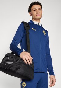 Giacca zip-up blu Puma con una finitura tessurizzata, caratterizzata da un logo giallo. Una borsa duffel nera con dettagli bianchi è tenuta con una tracolla nera.