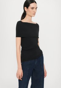 Zwarte off-shoulder top met een aansluitend ontwerp en geplooide textuur, gecombineerd met donkere spijkerbroeken met wijde pijpen. Glad materiaal met een subtiele glans.