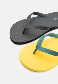 Paire de tongs en caoutchouc texturé, une noire avec des lanières noires et une jaune avec des lanières vertes, placées en angle sur un fond blanc.