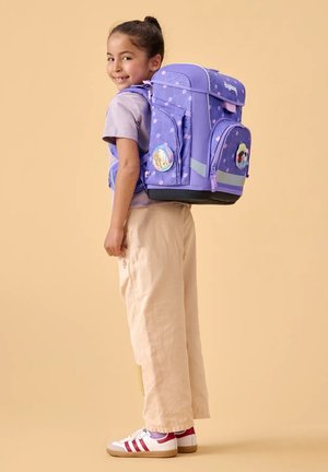 Lächelndes Mädchen mit dunklen Haaren im Dutt trägt beige Hose, lila Hemd und einen großen lila Rucksack mit Cartoon-Aufnähern, steht vor einem sandfarbenen Hintergrund.