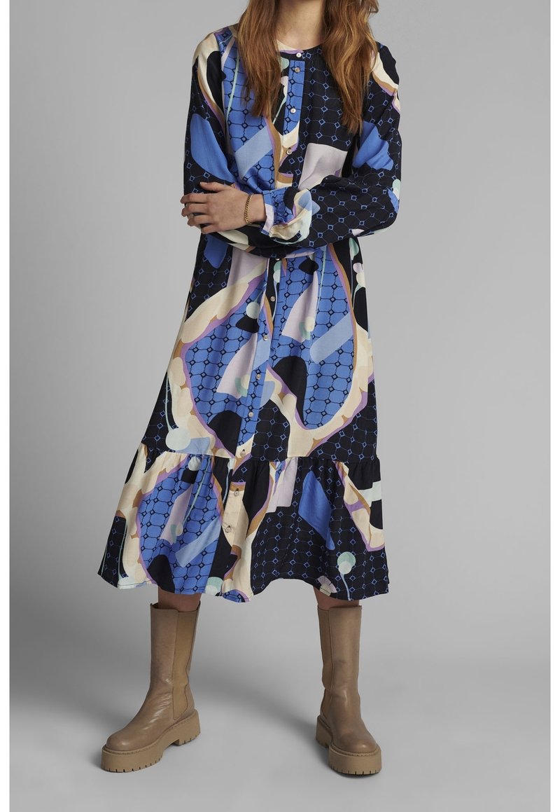 Femme portant une robe midi à manches longues avec des motifs abstraits bleus, noirs et beiges, associée à des bottes épaisses couleur beige montée jusqu'au milieu du mollet, debout les bras croisés.