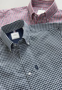 Se presentan dos camisas de botones, una a cuadros rojos y blancos, y otra a cuadros marinos y blancos, que destacan por su tela de algodón y diseño con cuello.